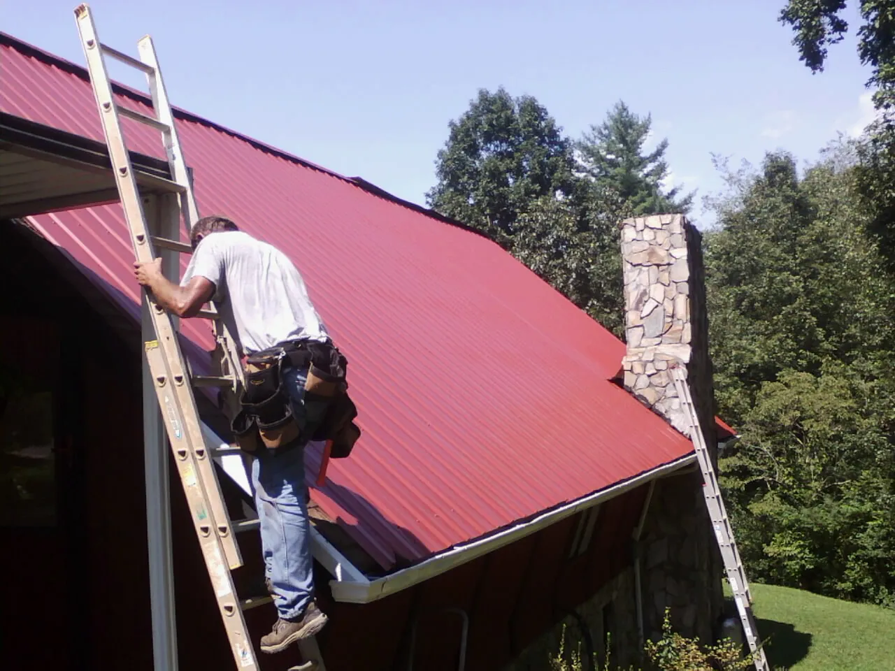 Roofer on a ladder beside a red metal roof and stone chimney — Soffit & Fascia Repair by Roofing Brothers in Napoleonville